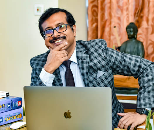 Dr. Pratim Sengupta is in a suit and tie, sitting at a desk with a laptop, and smiling at the front of the camera.