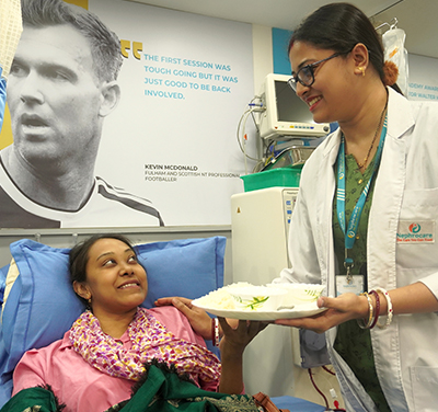 A nurse gives a plate of food to a woman patient lying on a hospital bed.