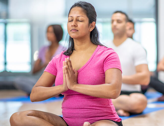 An Indian woman in a pink t-shirt is meditating by joining her hands and eyes close.